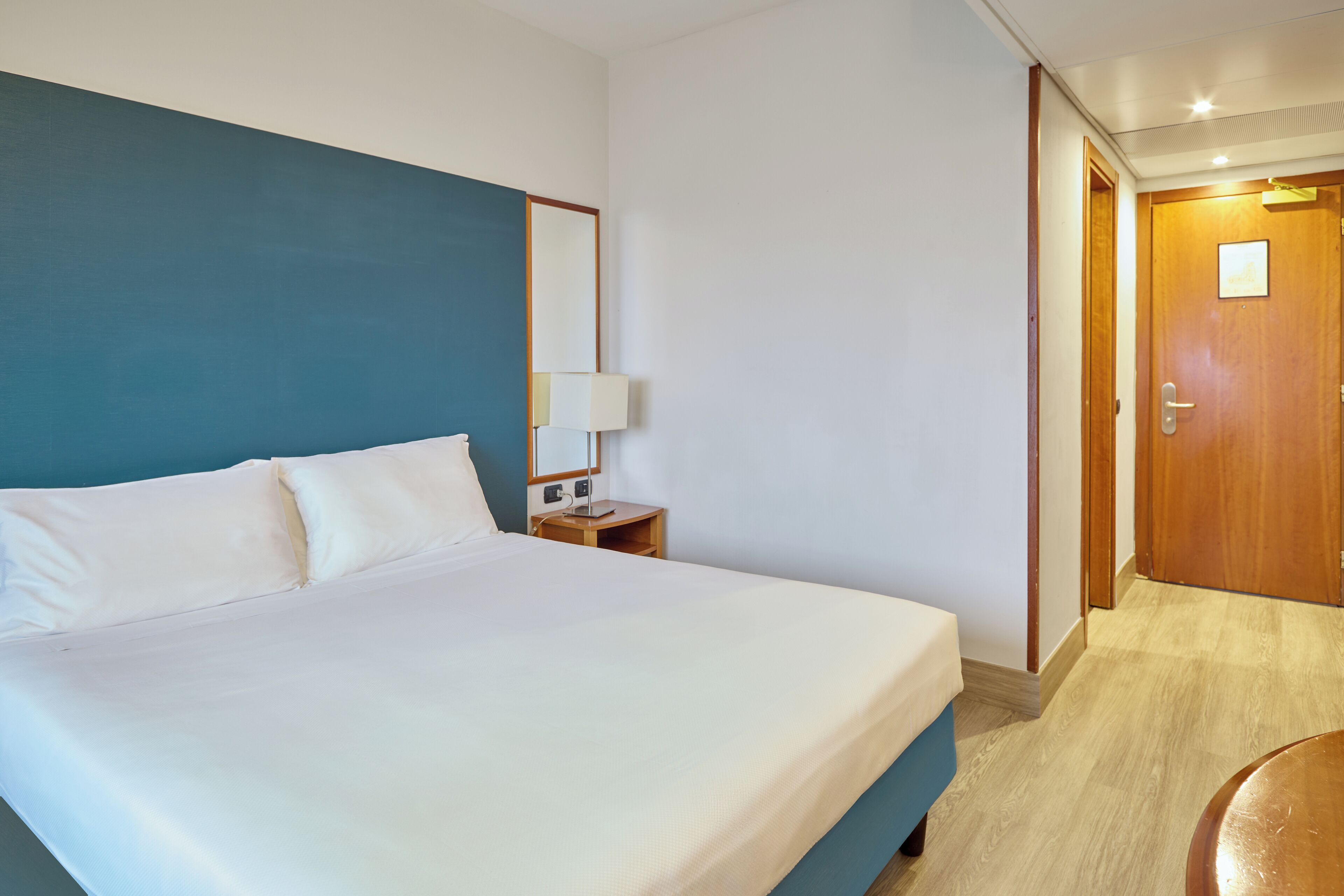 Una habitación de hotel con una cama grande, una mesita de noche con una lámpara y una puerta de madera.
