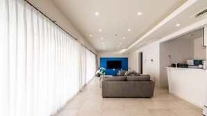 Una sala moderna con un sofá gris, un televisor de pantalla plana montado en una pared azul y una zona de cocina con microondas y cafetera.