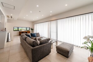 A modern living room with a sofa, ottoman, and a glass coffee table. There is a kitchen area with a refrigerator and a TV. - Villa Alyssum (Miyakojima)