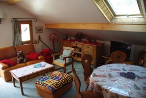 Uma sala de estar aconchegante com um sofá, poltrona e pufe, uma mesa de madeira com um colcha, uma televisão e uma janela com vista.
