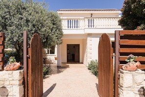 Una villa con entrada pavimentada, un portón de madera y un balcón con barandilla blanca.