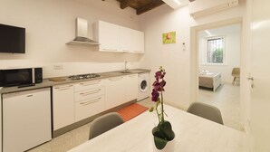 A modern kitchen with white cabinets, a microwave, and a gas stove. There is a dining table with chairs and a potted orchid. A washing machine is visible in the background.