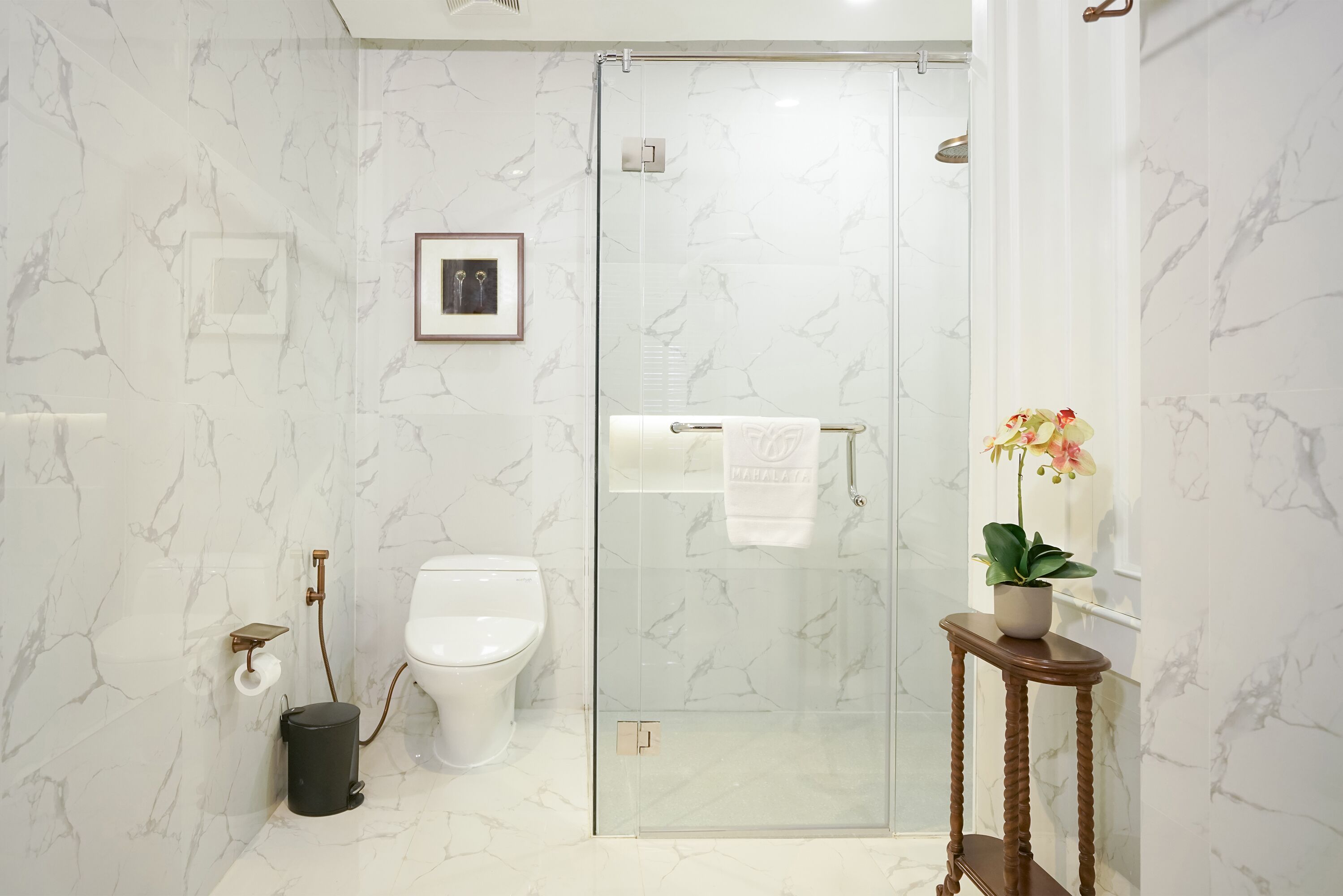 A modern bathroom with a glass shower enclosure, a toilet, a marble wall, a framed picture, and a potted plant.