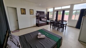 A hotel room with a bunk bed, a single bed, a dining table with chairs, and large windows.