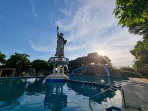 Une statue se trouve dans un espace aménagé autour d’une piscine, avec un ciel dégagé et des arbres en arrière-plan.