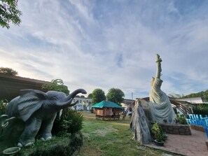 Un jardín con una estatua de un elefante y una estatua de una figura sosteniendo un báculo. Hay plantas en macetas y una estructura de madera al fondo.