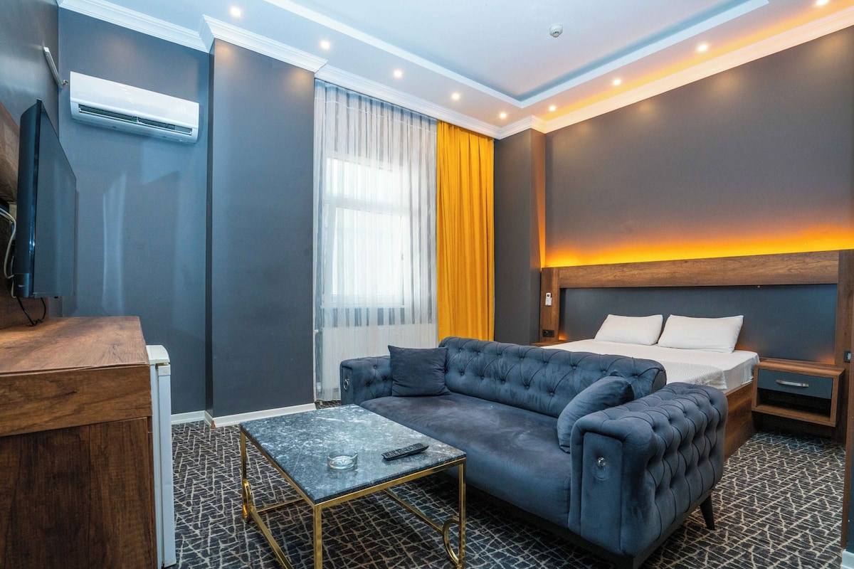 Une chambre d’hôtel moderne avec un canapé gris, une petite table, un lit avec une tête de lit en bois et un appareil de climatisation.