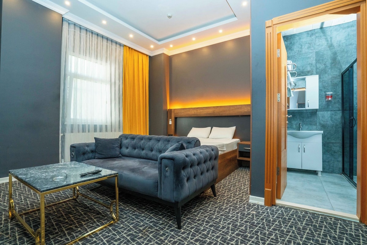 Une chambre d’hôtel moderne avec un canapé gris, une petite table, un lit avec une tête de lit en bois et une salle de bain visible à travers une porte ouverte.