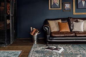 Un salon à l’ambiance sombre, avec un canapé en cuir, un seau en métal rempli de bois de chauffage et un tapis à motifs.