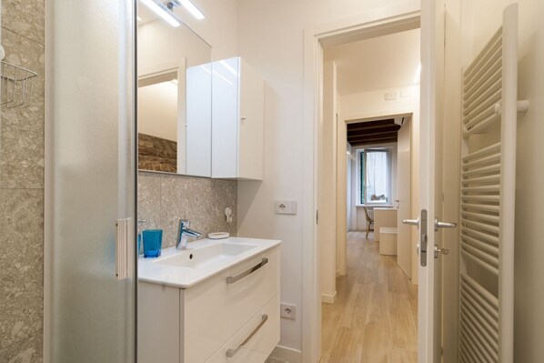 Un baño moderno con un tocador blanco, un espejo y un lavabo. Se observa una zona de ducha a la izquierda y una puerta que da a otra habitación.