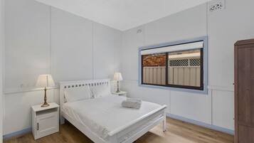 A bedroom with a bed, two bedside tables with lamps, a window with a view of a fence, and a wardrobe.