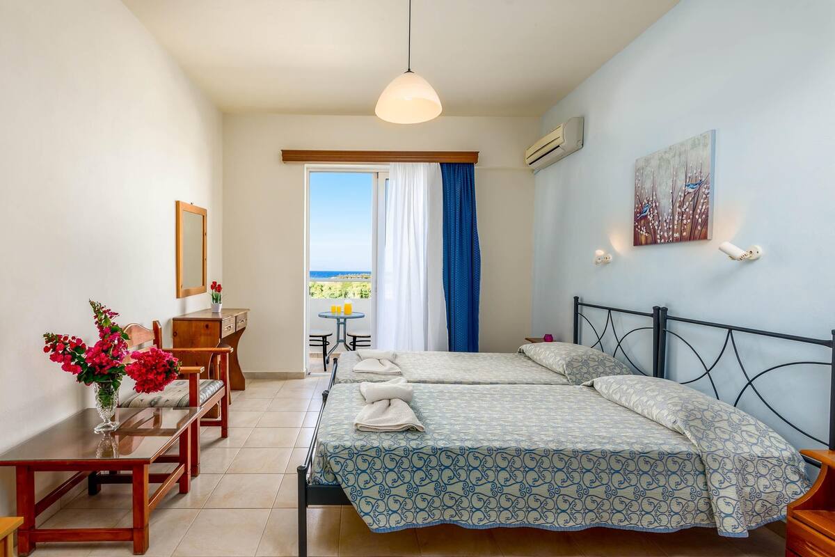Habitación de hotel con dos camas, una mesa de comedor, una silla, una mesita con un florero y vista al mar a través de una ventana.