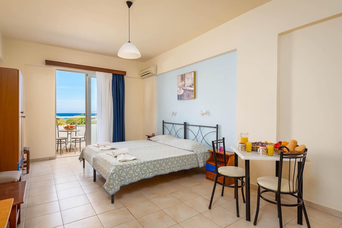 Habitación de hotel con cama, una mesa de comedor con sillas, un balcón con vista al mar y un cuadro en la pared.