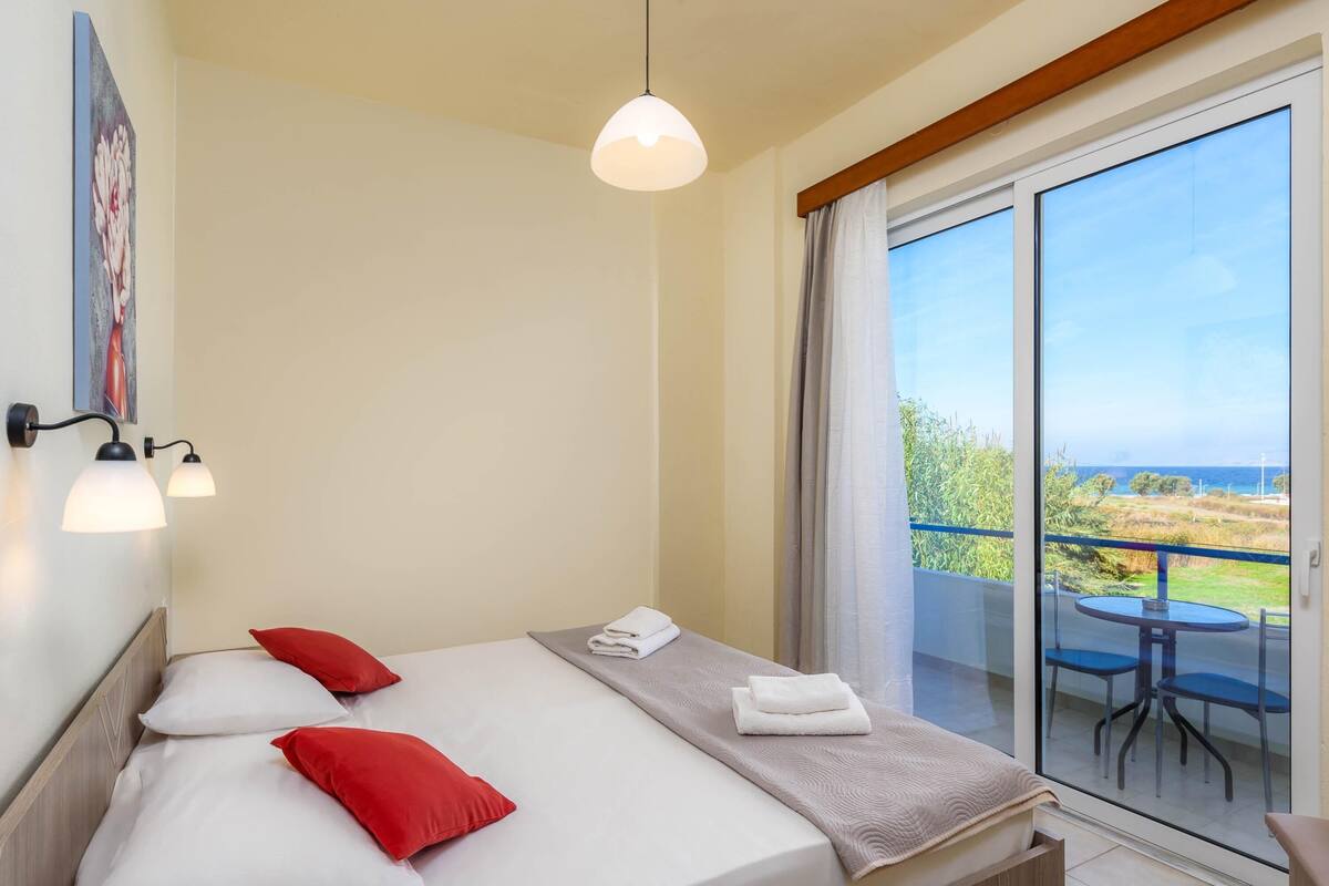 Habitación de hotel con cama, almohadas rojas, manta blanca, toalla y balcón con mesa y sillas.