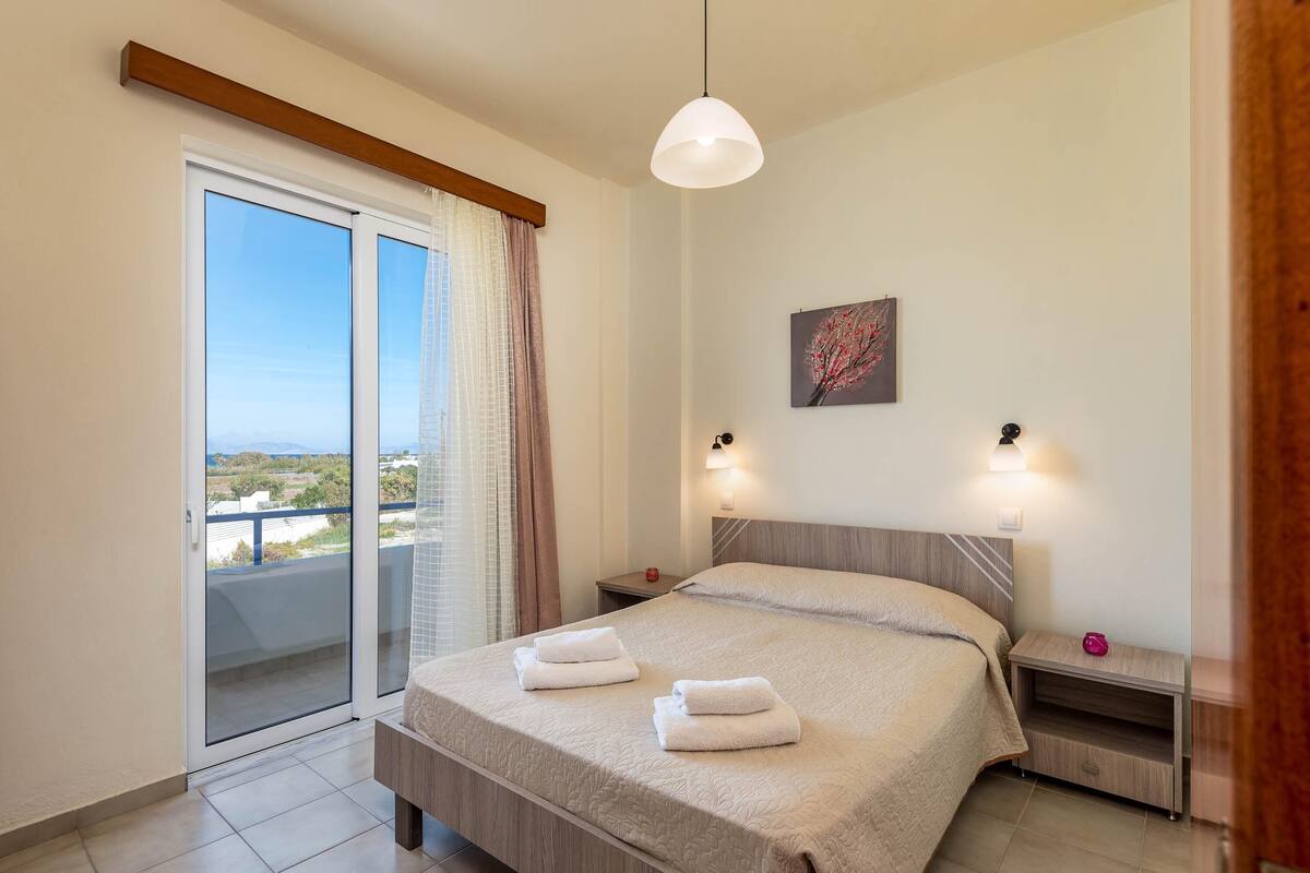 Un dormitorio con una cama, dos mesitas de noche, una lámpara de noche, una luz de techo, una ventana con cortinas y vistas al exterior.