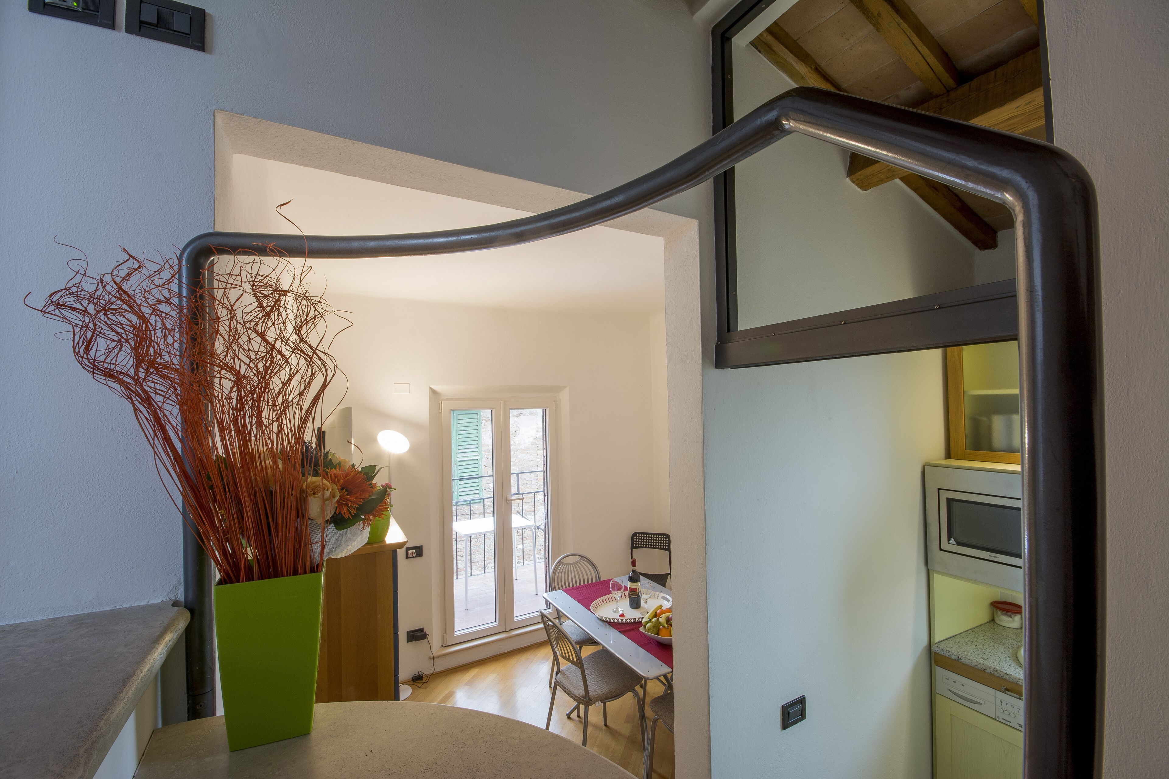 A staircase with a green vase of dried plants, a dining area with a table and chairs, and a kitchen area with a microwave.