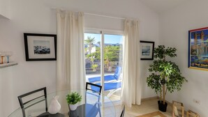 A modern room with a glass table, chairs, a potted plant, and a view of a pool and greenery outside. - C2, en pequeño complejo con piscina comunitaria a 100m del mar (Charco del Palo)