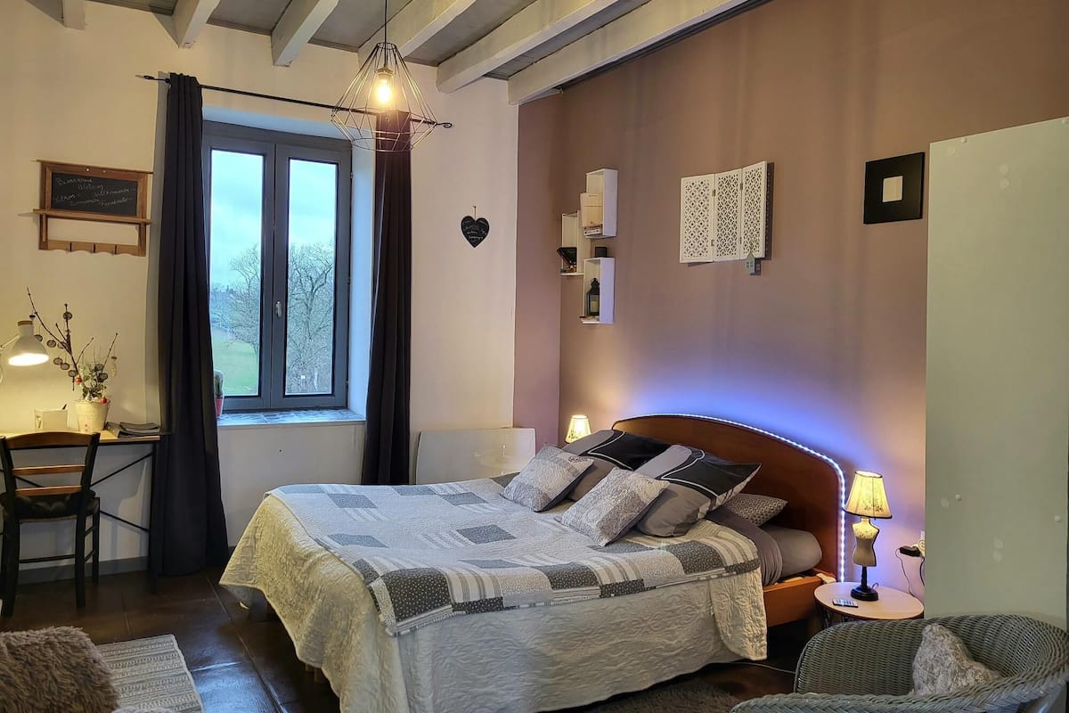 Un dormitorio con cama, mesita de noche, lámpara, silla y ventana con cortinas.