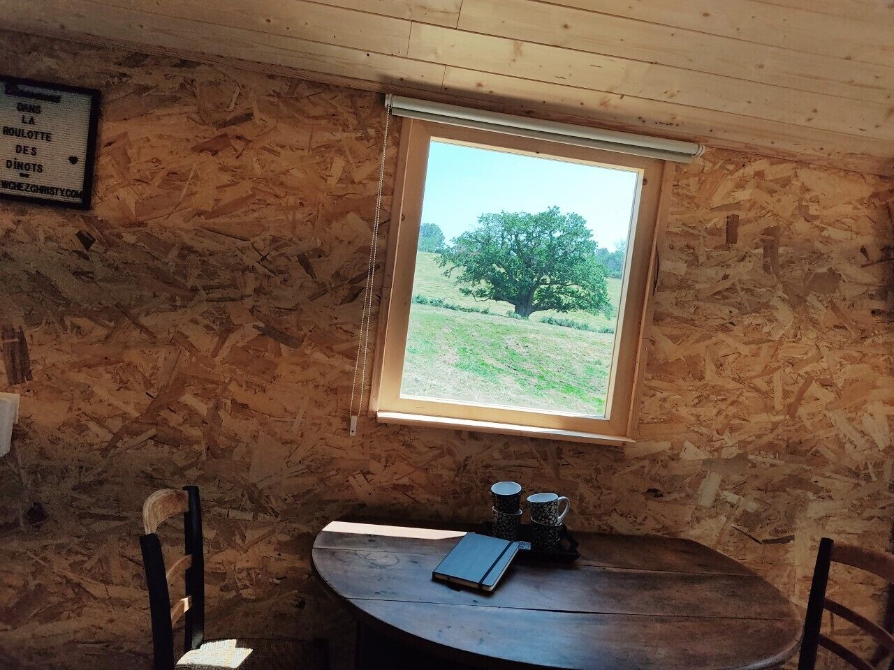 A rustic room with a wooden wall, a small table with a notebook, two cups, and a window with a view of a tree and field.