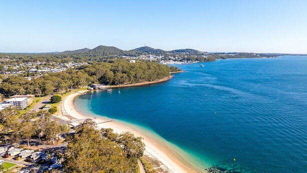 An aerial view of a coastal area with a beach, clear blue waters, and a forested hill in the background. - Veronica Court 1, 4 Weatherly Cl - Aircon (Nelson Bay)