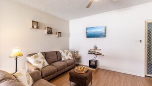 A living room with a sofa, a coffee table with pastries, a TV mounted on the wall, and a small side table with a lamp. - Veronica Court 1, 4 Weatherly Cl - Aircon (Nelson Bay)