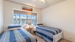 A bedroom with two beds, a window with blinds, a ceiling fan, and a painting on the wall.