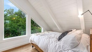 Ein Schlafzimmer mit schrägen Wänden, großen Fenstern, einem Bettgestell aus Holz und einem Nachttisch mit Lampe.