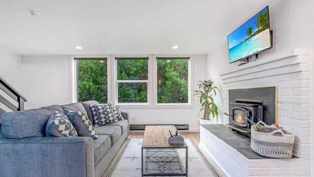 Ein modernes Wohnzimmer mit grauem Sofa, Kamin, Wand-Fernseher und großen Fenstern mit Blick auf Grünflächen.