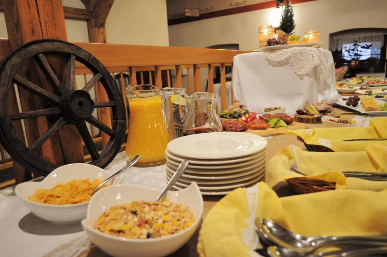 A breakfast buffet with various dishes, a wooden chair, and a table with stacked plates and a pitcher of orange juice.