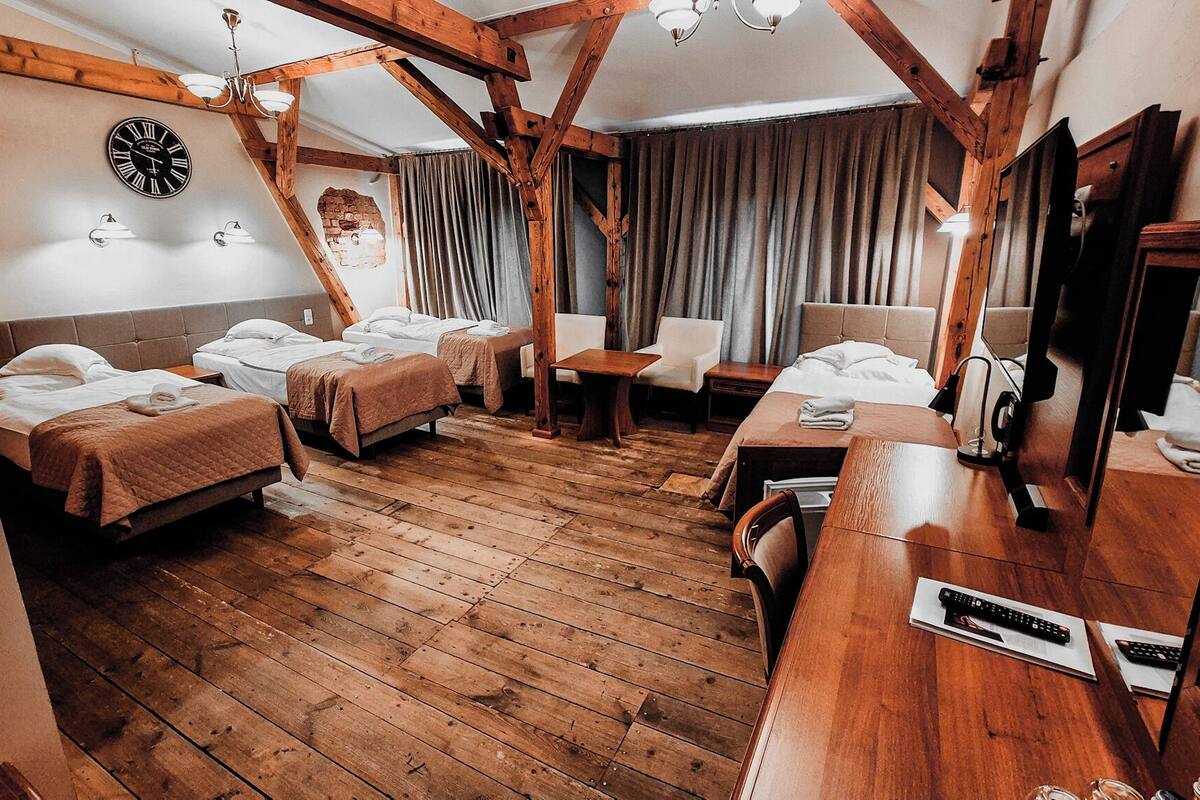 Une chambre d’hôtel avec un plancher en bois, un grand lit, deux petits lits, une télévision, un bureau et une horloge au mur.