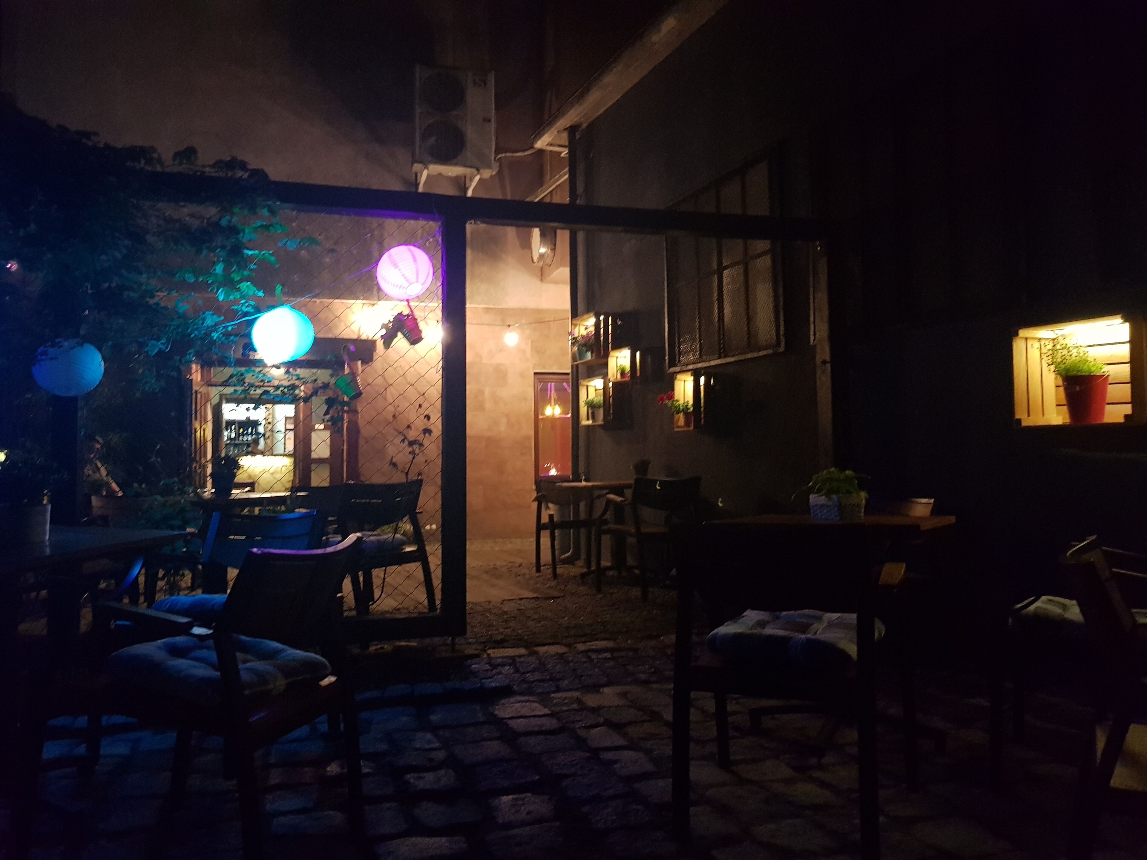An outdoor seating area with tables and chairs, a brick pathway, and a building entrance with a lit window.