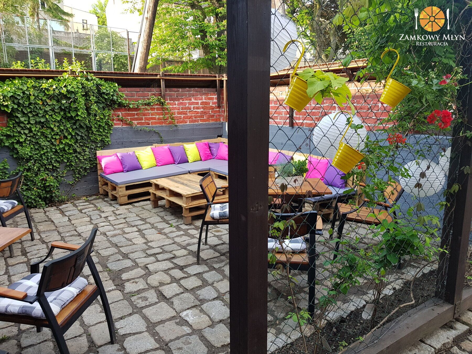 An outdoor seating area with wooden tables and chairs, a cushioned bench with colorful cushions, and a brick wall with greenery.