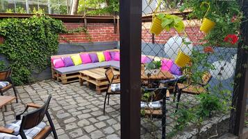 An outdoor seating area with wooden tables and chairs, a cushioned bench with colorful cushions, and a brick wall with greenery.