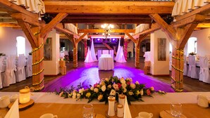 Un espacio para recepción de bodas con techo de madera, mesas dispuestas con manteles blancos y sillas, y un arreglo floral en el suelo.