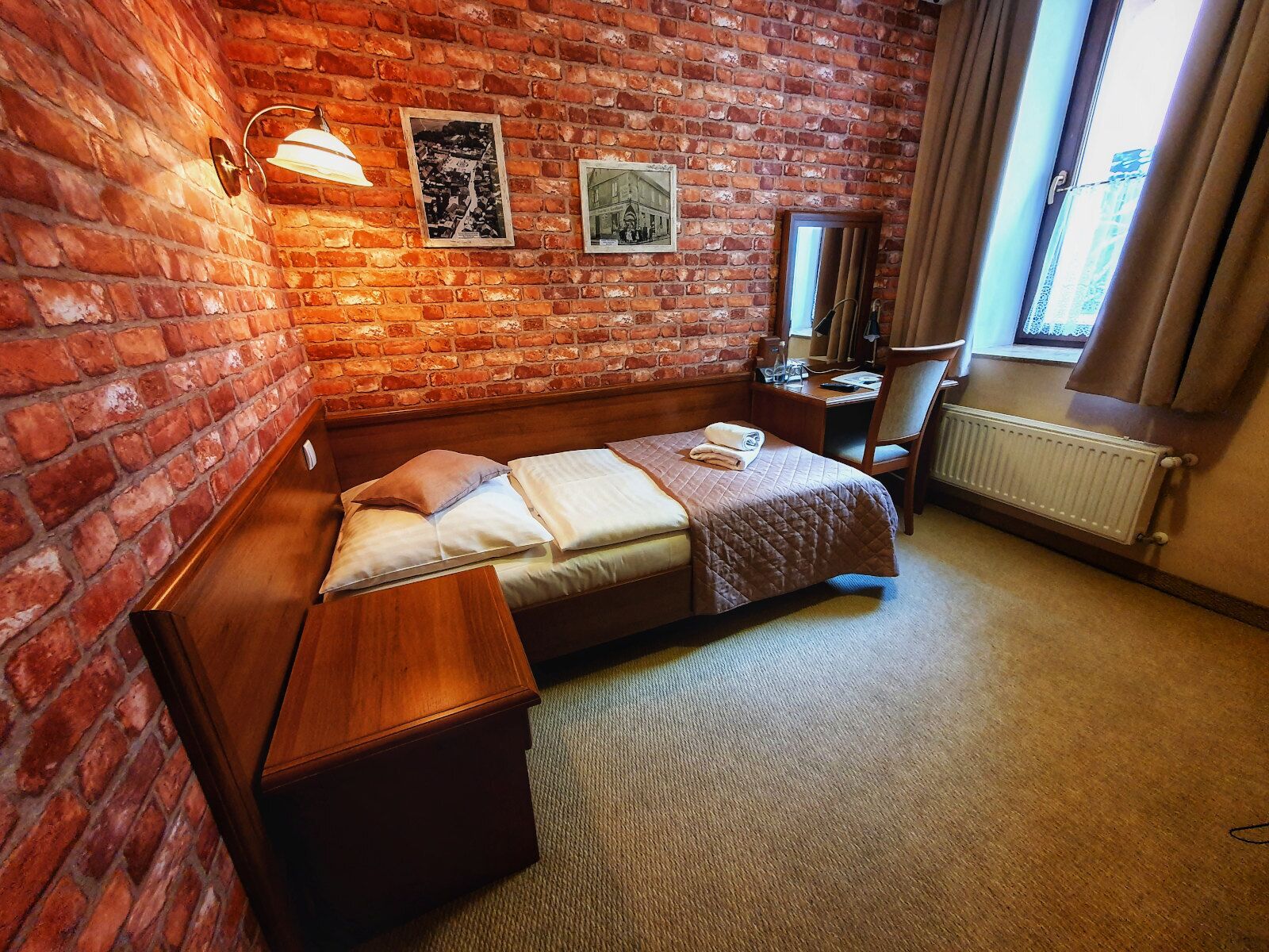 A hotel room with a brick wall, a bed with a quilt, a nightstand, a desk, a chair, and a window with curtains.