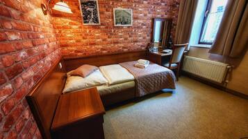 A hotel room with a brick wall, a bed with a quilt, a nightstand, a desk, a chair, and a window with curtains.