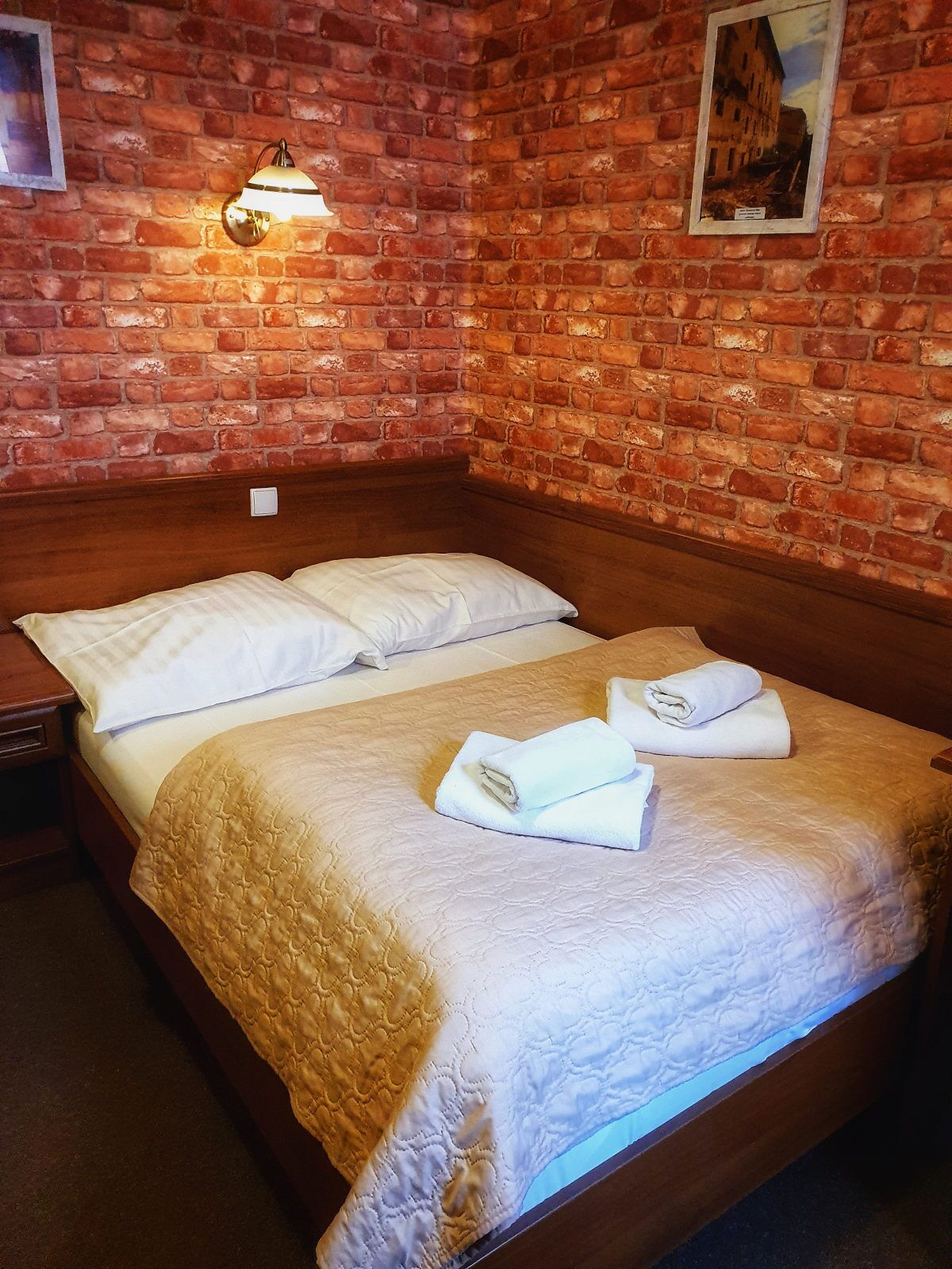 A double bed with white linens and a quilted cover, a nightstand, a wall-mounted lamp, and a framed picture on a brick wall.