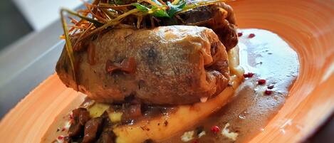 A dish with a roasted meat centerpiece, garnished with herbs, served on a plate with a sauce and vegetables.