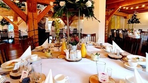 Un comedor con vigas de madera y una araña, mesas dispuestas con manteles blancos, platos y copas.