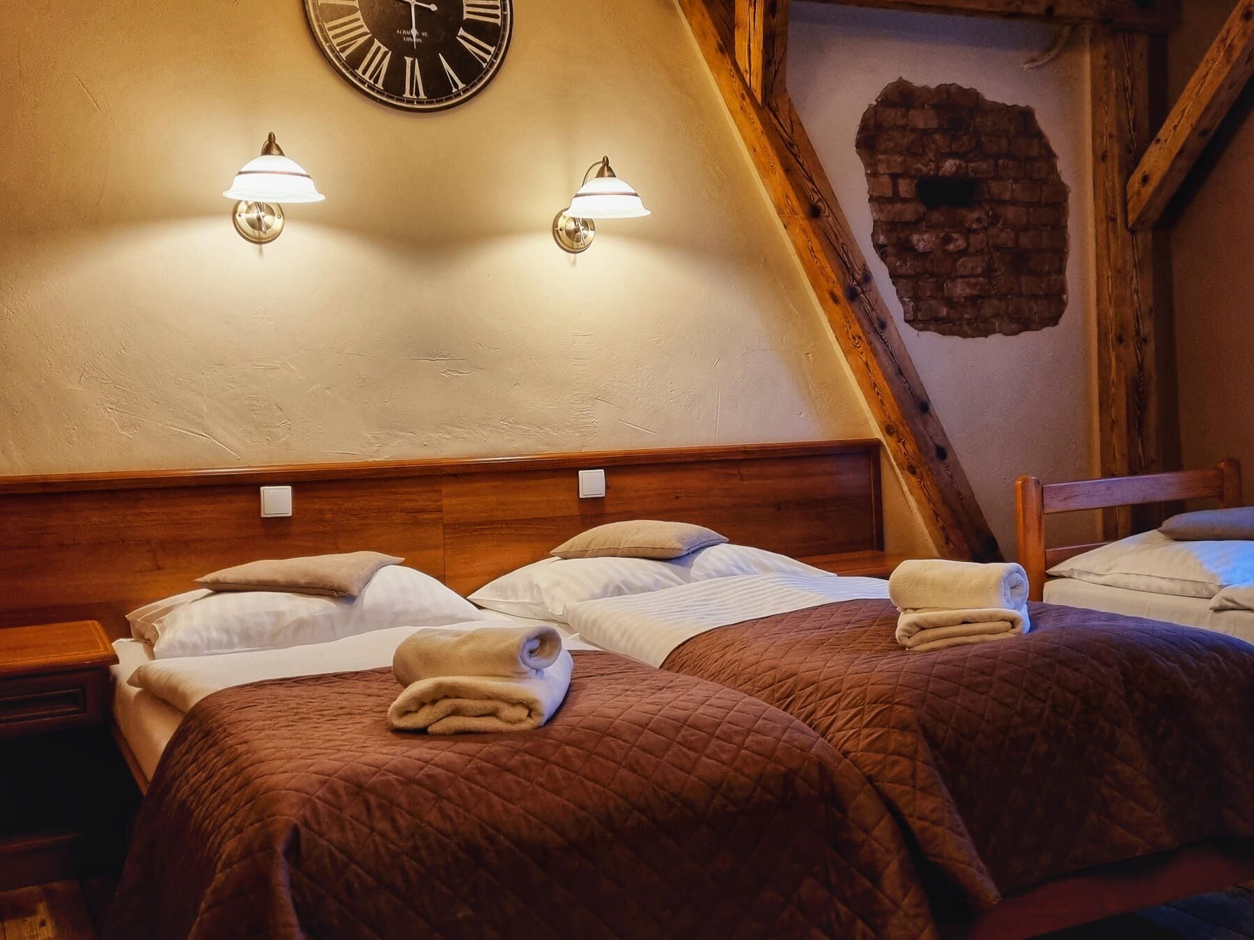 A room with two beds, each with a wooden headboard, and a brick wall with a clock above.
