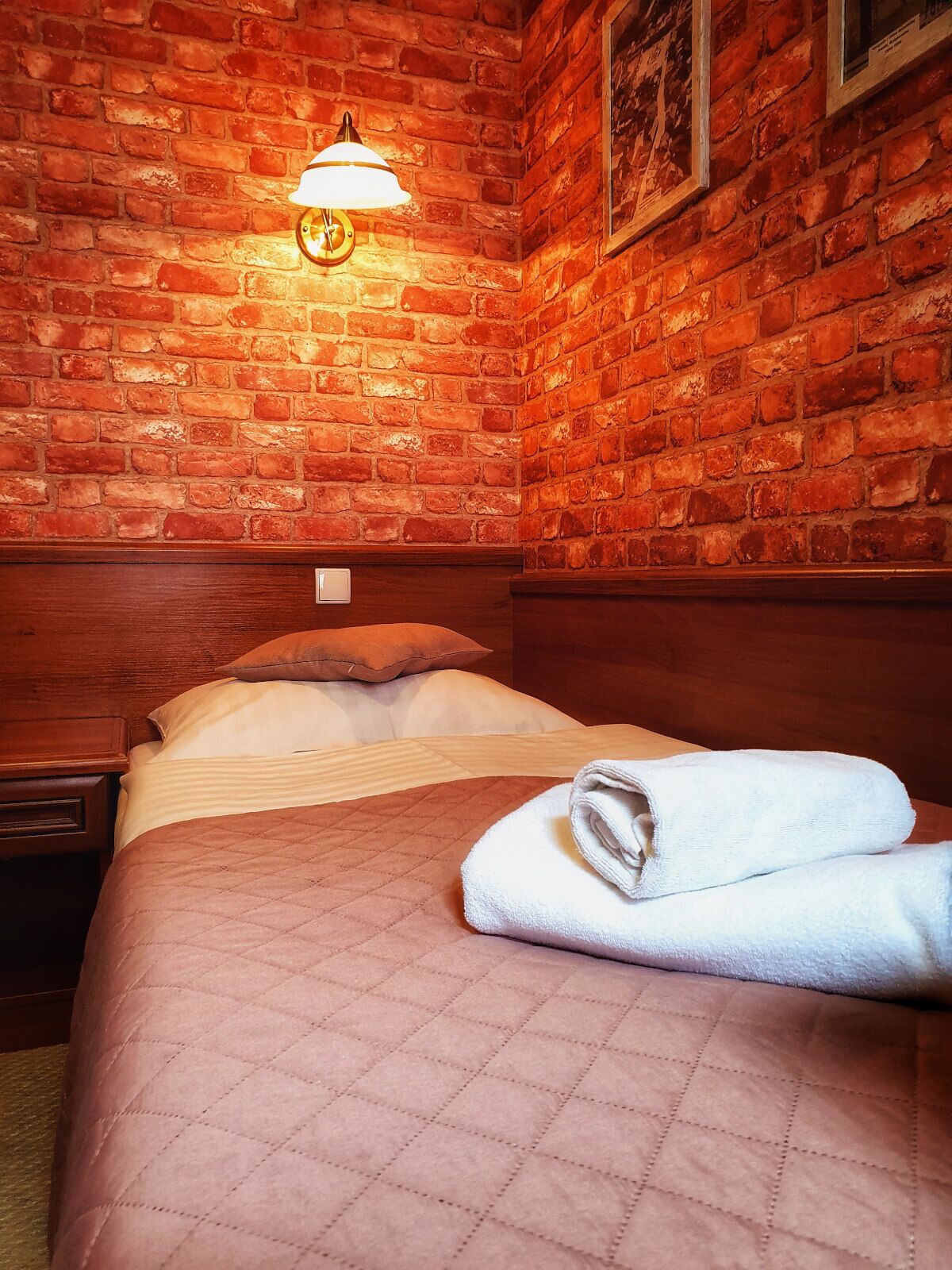 A hotel room with a brick wall, a bed with a quilted cover, a white towel, and a nightstand.