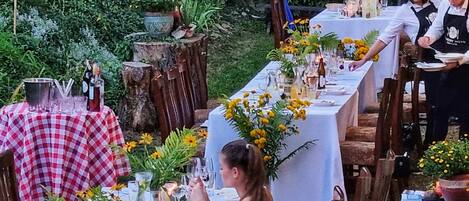 Un área de comedor al aire libre con una larga mesa dispuesta para una comida, rodeada de follaje y elementos de madera rústica.