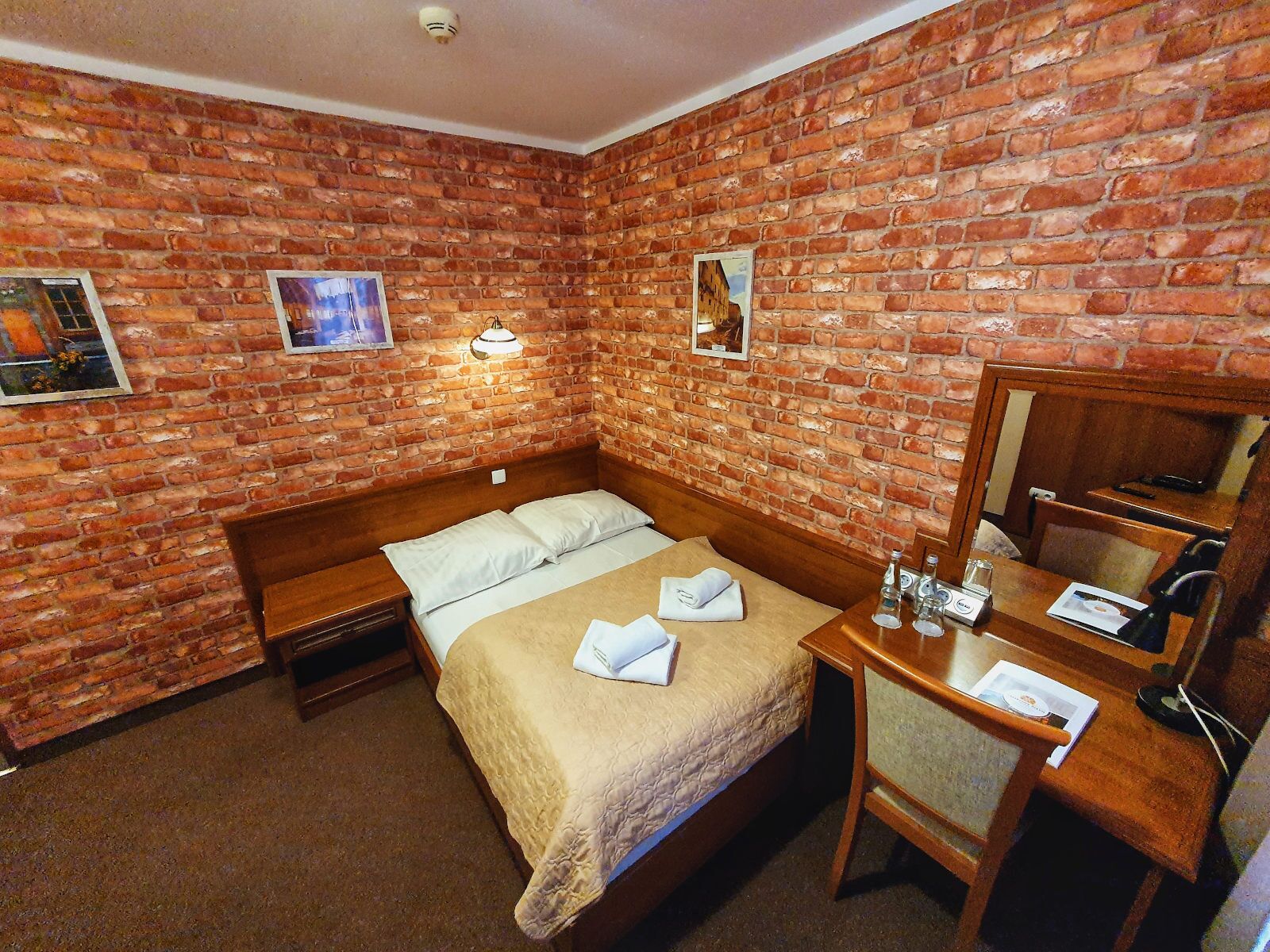 Une pièce avec un mur en briques, un lit recouvert de draps blancs, une table de chevet avec un miroir et un petit bureau avec une chaise.