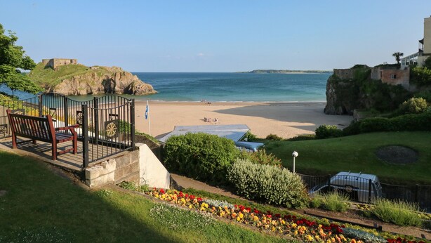 Vista costeira com praia, um edifício histórico e um jardim com flores coloridas.