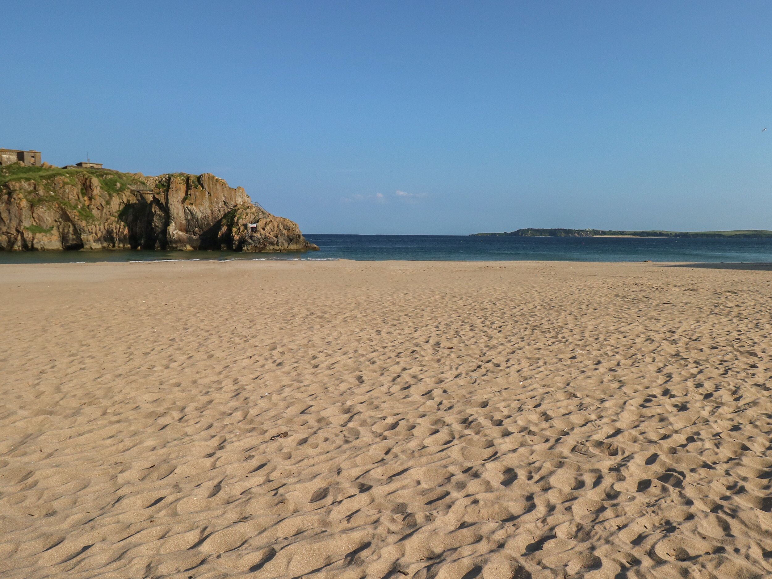 A sandy beach with footprints leading to the water, a rocky cliff, and a clear sky.