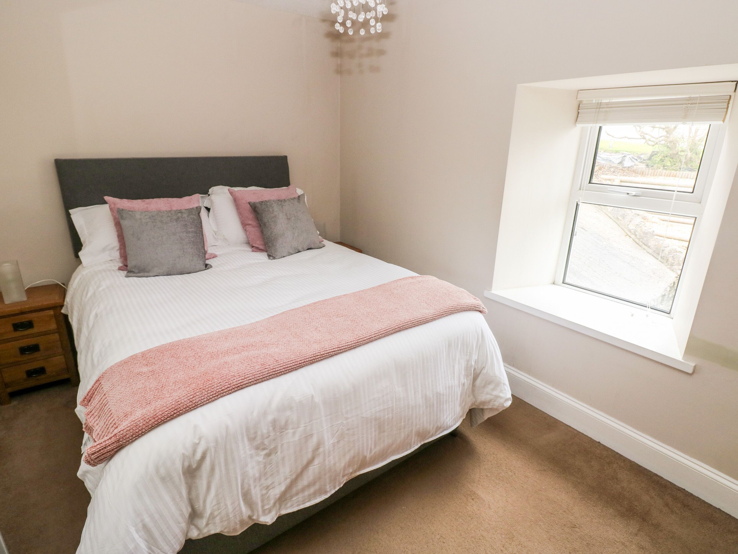 A neatly made bed with a white and pink bedspread, a wooden nightstand with a lamp, and a window with blinds.