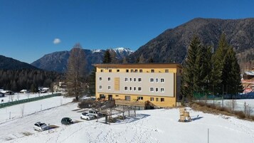 Un hotel de varios pisos con techo plano, situado en una zona nevada con montañas al fondo.
