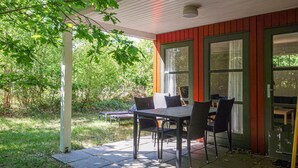 Een terras buiten met een tafel en stoelen, omgeven door groen en een gebouw met grote ramen.