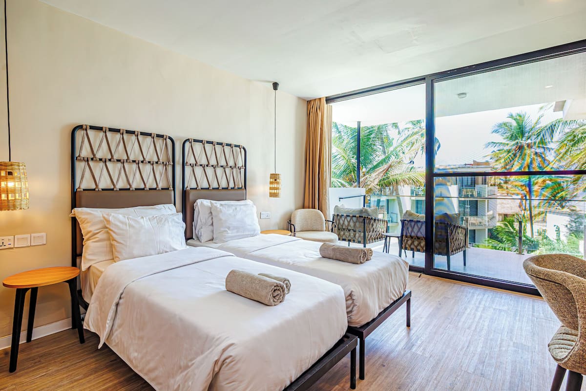 Habitación de hotel con dos camas, un balcón con una silla y vista a palmeras.