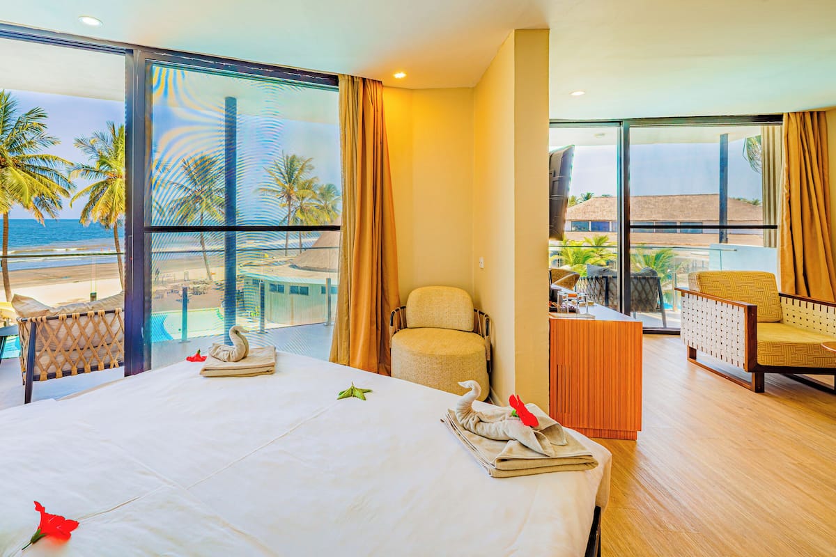 Habitación de hotel con una cama grande, una silla, una mesita y vistas a la playa y al océano a través de ventanas amplias.